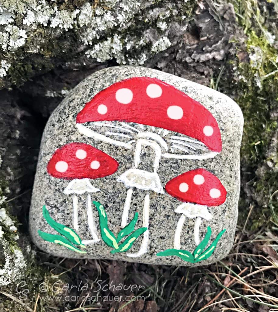 3 mushrooms with red caps and white dots, painted on a speckled rock, sitting on a tree bark background